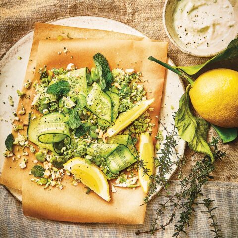 Fresh green coloured vegetable salad with shaved cucumber ribbons, herbs and chopped nuts on parchment paper, served with lemon wedges and creamy dressing.