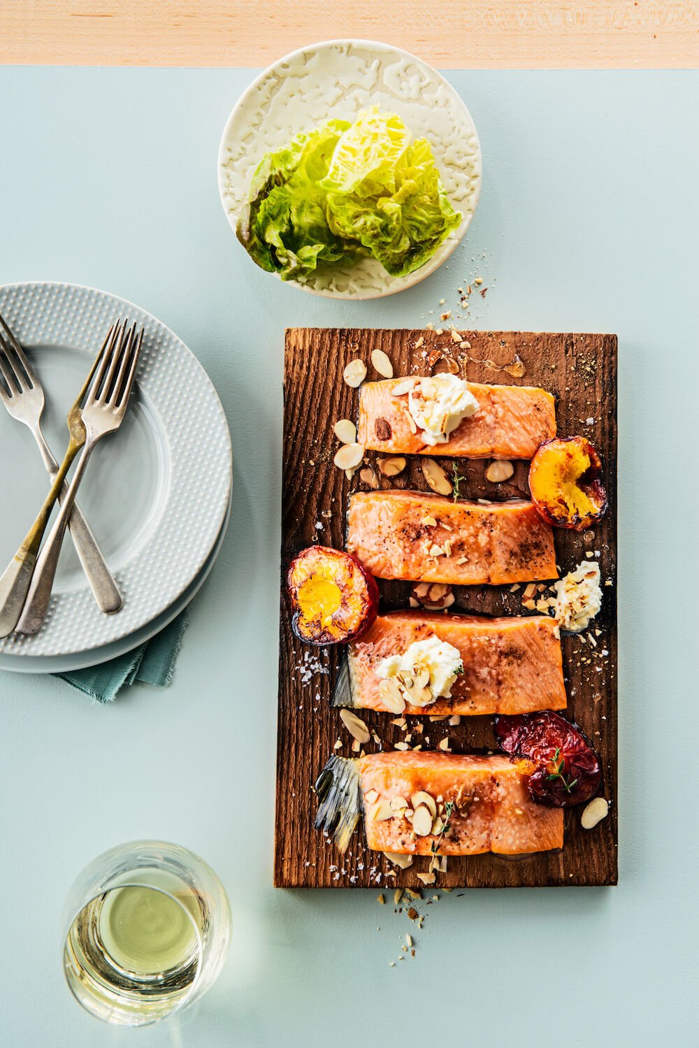 Four salmon fillets on a cutting board with plates, salad and wine on the table nearby.