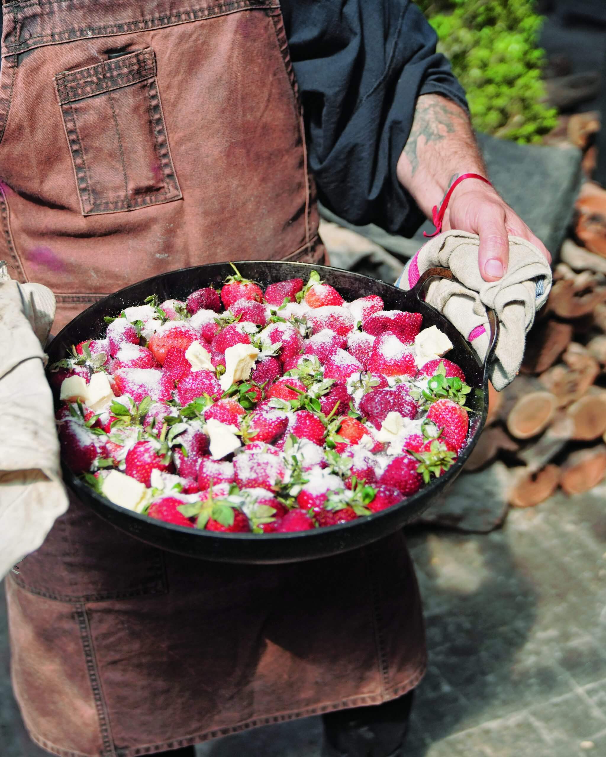 A man holding a cast-iron dish filled with roasted strawberries, ricotta and mint