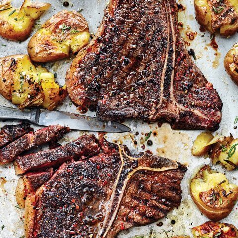 Two T-bone steaks and smashed potatoes on a pan with knife.