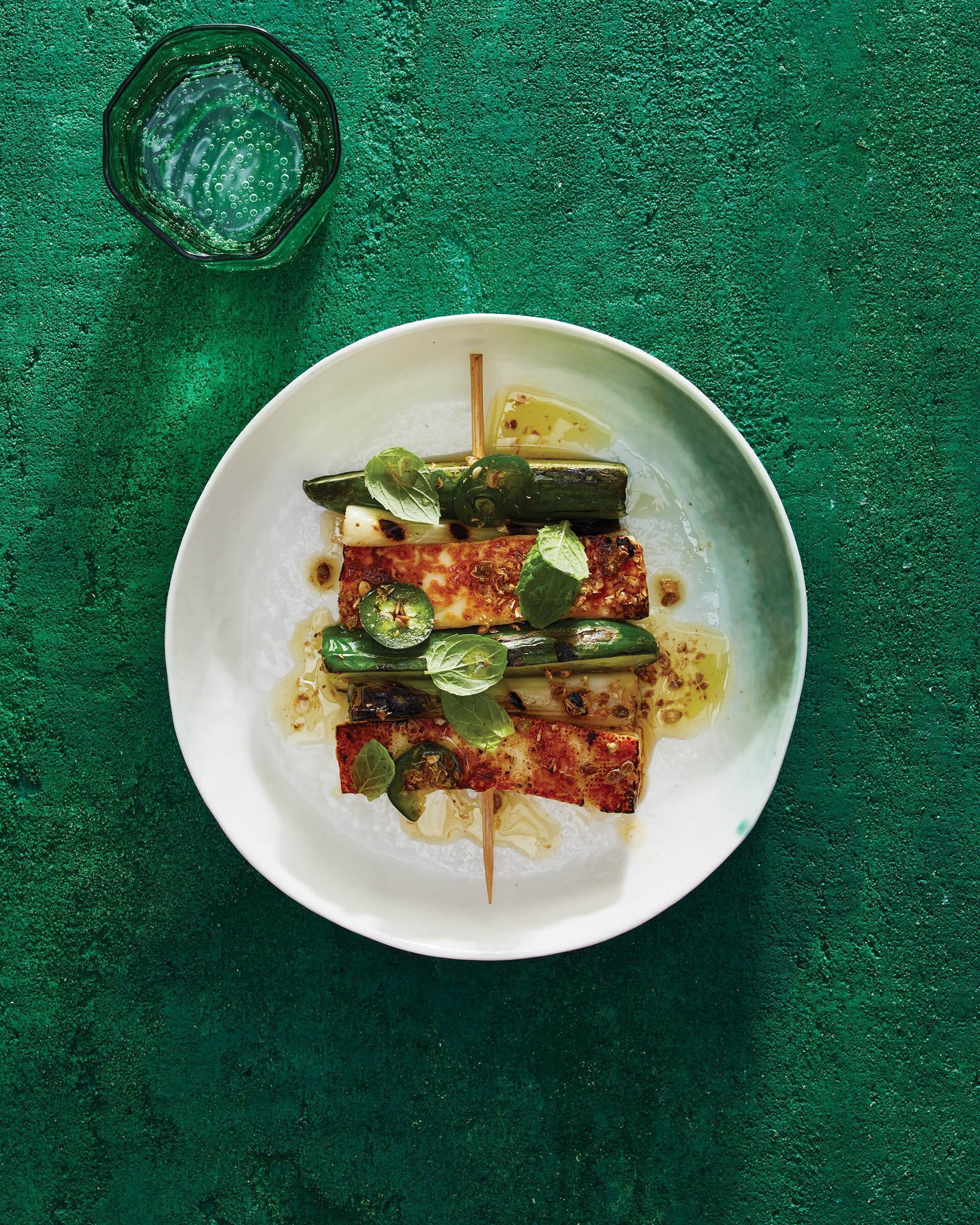 Skewered cucumbers and halloumi cheese on a white dish against a bright green table.
