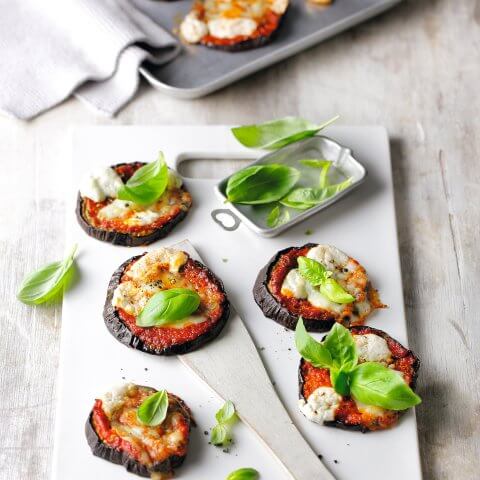 Eggplant rounds with toppings and basil garnish on a cutting board with a baking ban in the background.