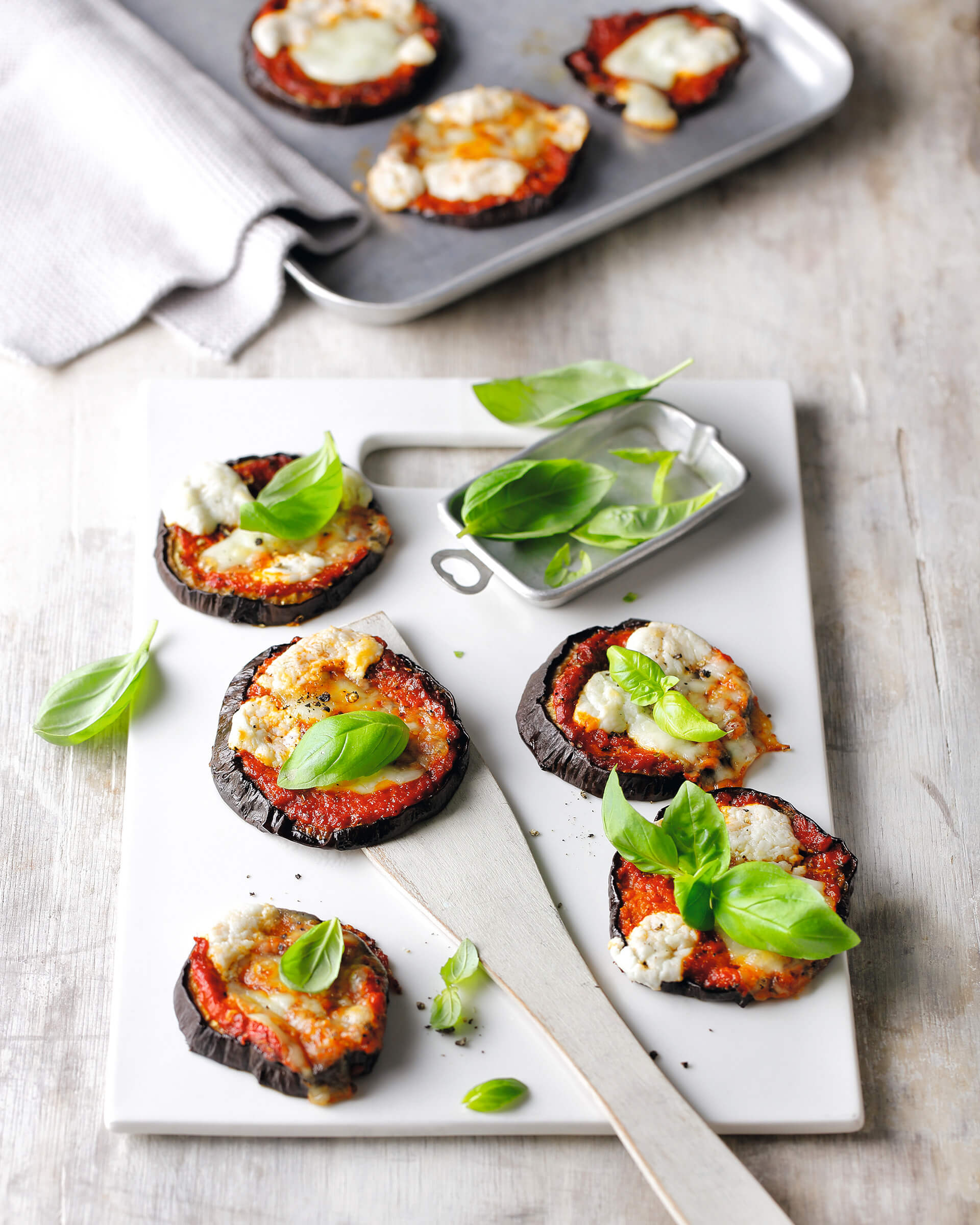 Eggplant rounds with toppings and basil garnish on a cutting board with a baking ban in the background.