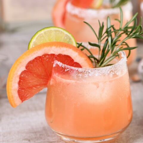 Two short glasses with a pink drink and grapefruit, lime and sage garnish.
