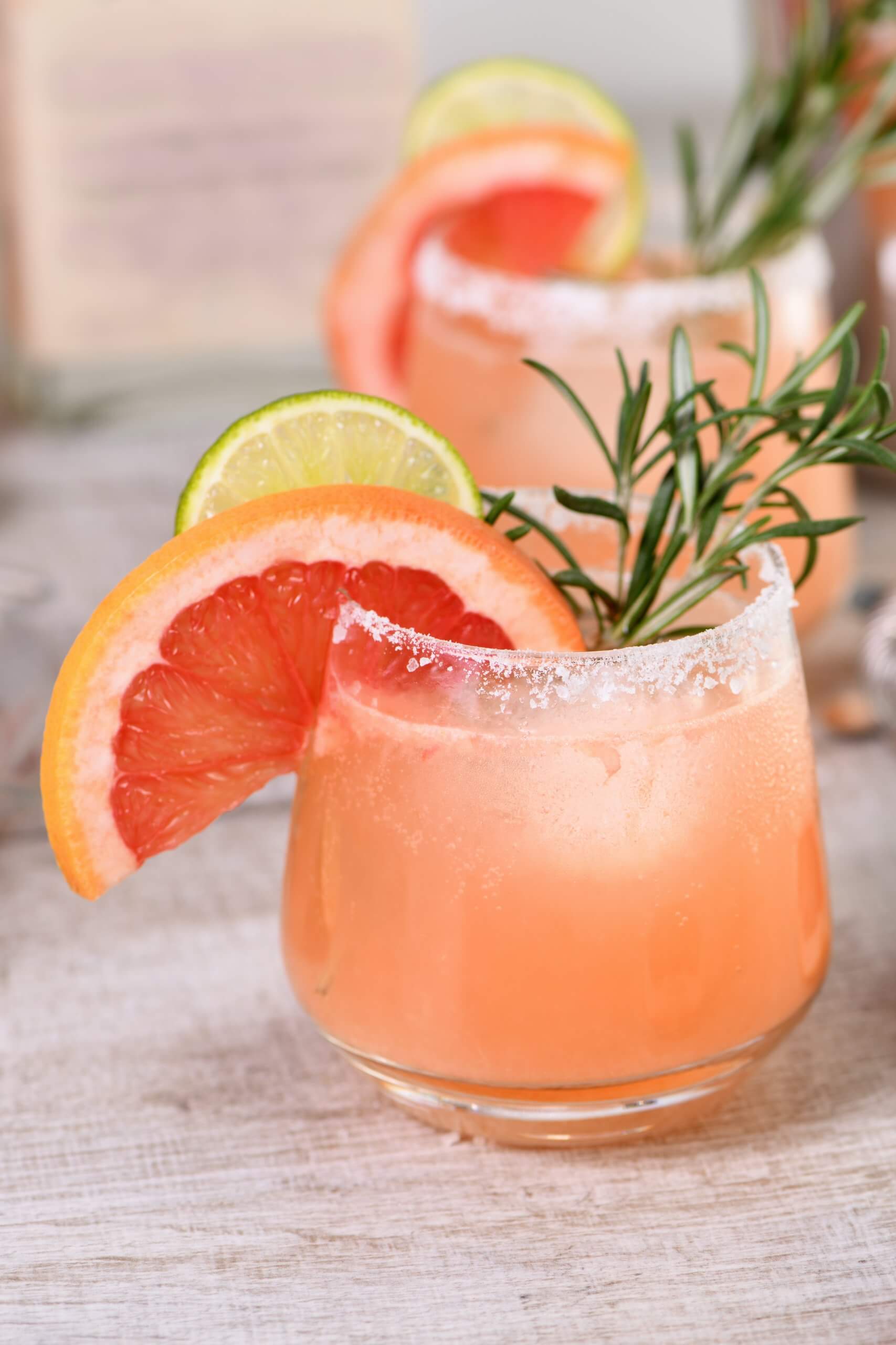 Two short glasses with a pink drink and grapefruit, lime and sage garnish.