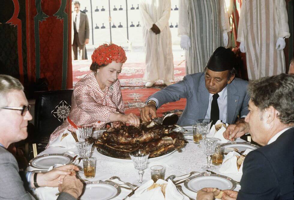 Queen Elizabeth reaches towards a platter of meat at a dining table with three men
