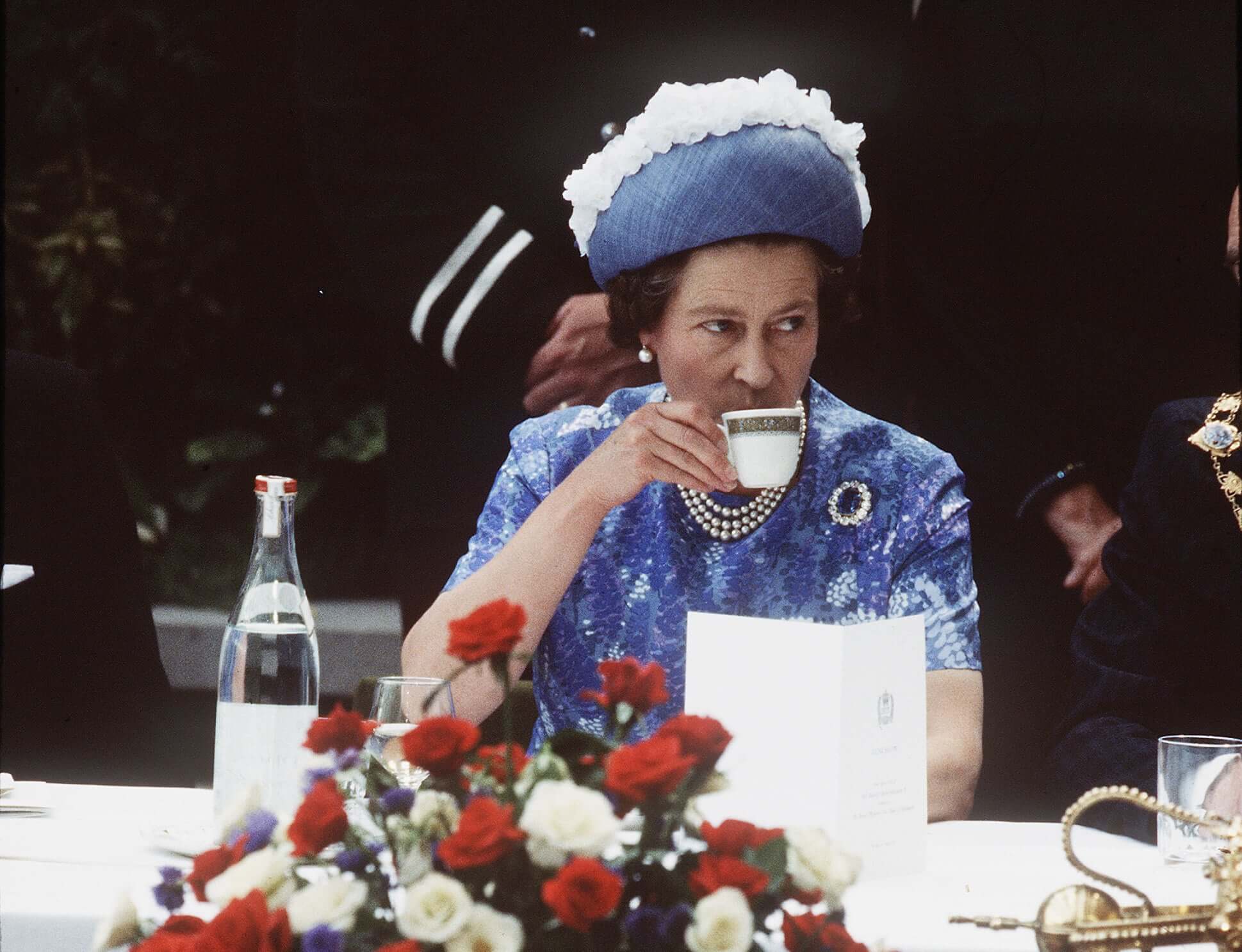 Queen Elizabeth, dressed in blue, sips from a teacup