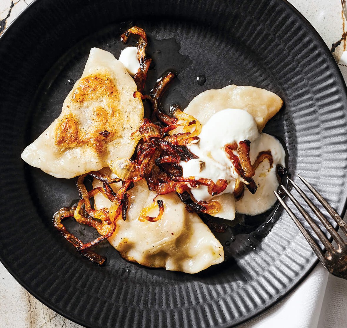 A black plate on a marble surface with pierogi, sour cream, bacon, onion and a fork
