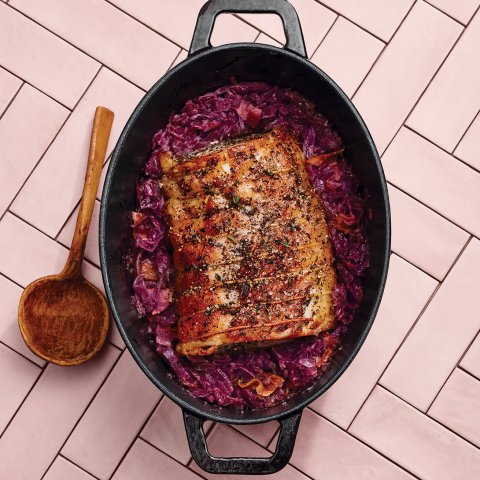 A cast iron dish with pork and cabbage and a wooden spoon on a pink countertop