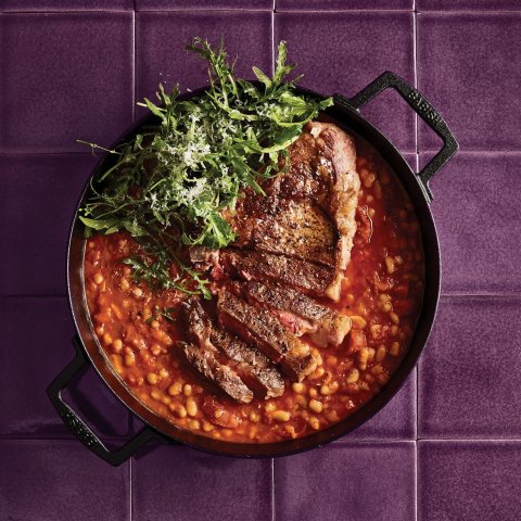 A cast iron pot with steak, beans and leafy greens on a purple countertop