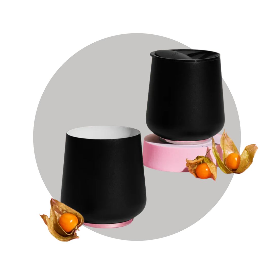 Two black wine tumblers on a pink pedestal with goldenberries on a decorative gray circle background. 