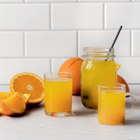 An orange drink in a mason jar and glasses surrounded by sliced oranges.