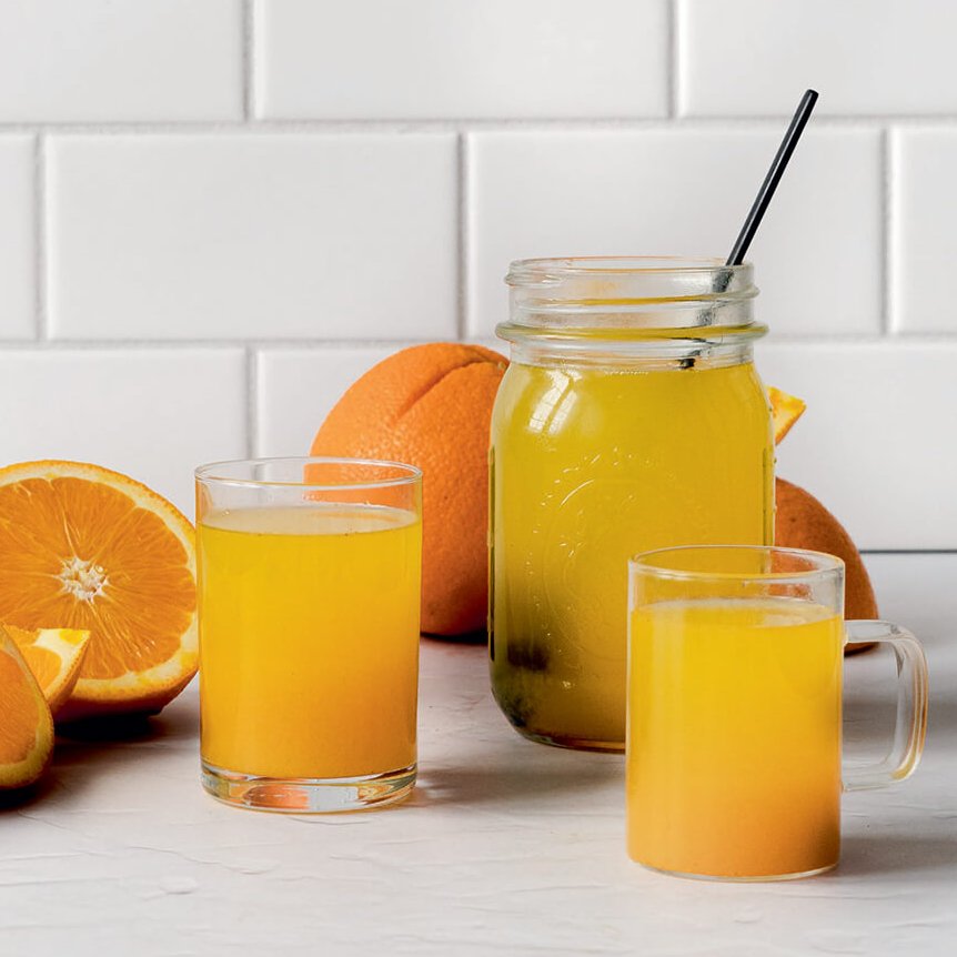An orange drink in a mason jar and glasses surrounded by sliced oranges.