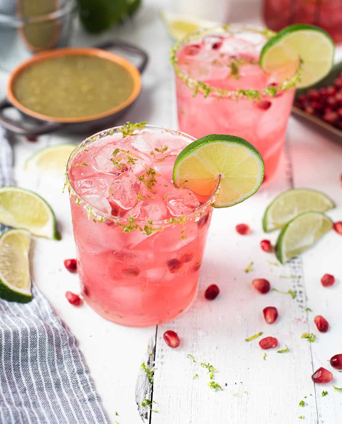 Glasses with a pink cocktail and lime garnish surrounded by pomegranate seeds.