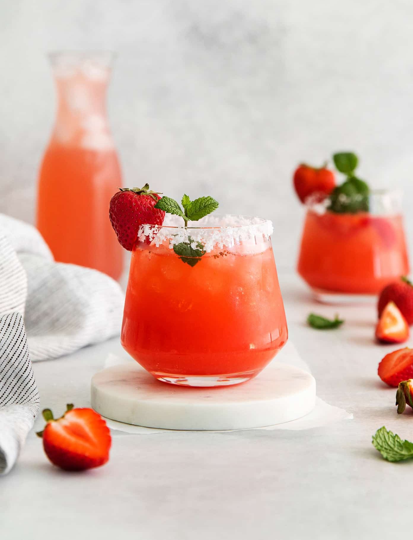 Glasses of a red drink garnished with strawberries and a pitcher in the background.