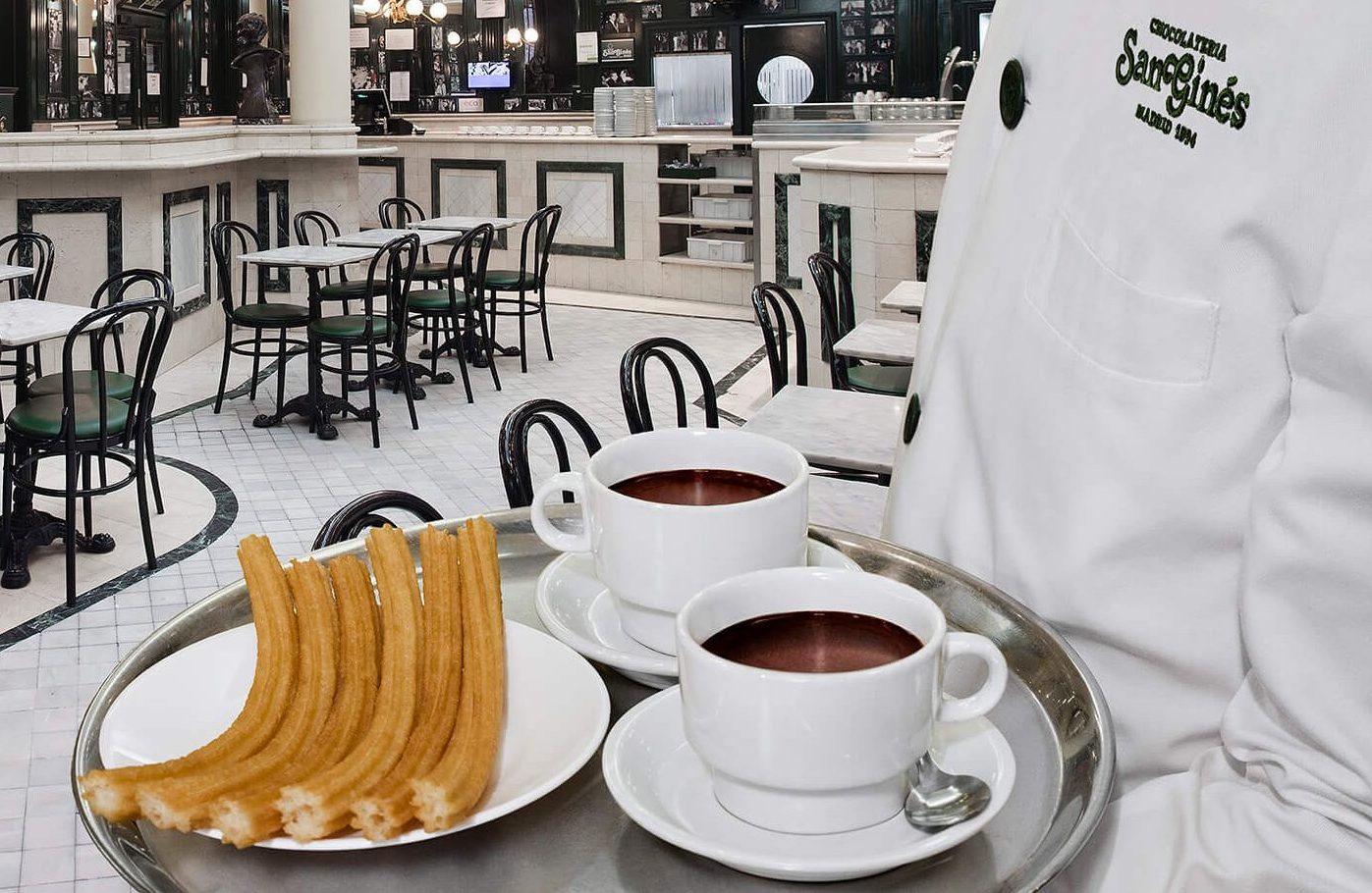 A server in white holds a tray with hot chocolate and churros, with an empty restaurant dining room in the background.