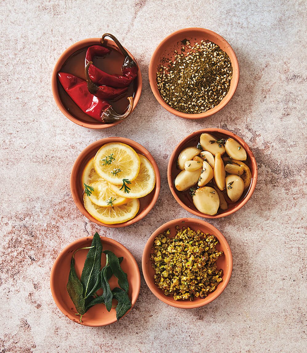Six terracotta bowls with various spices and garnishes on a light stone surface.