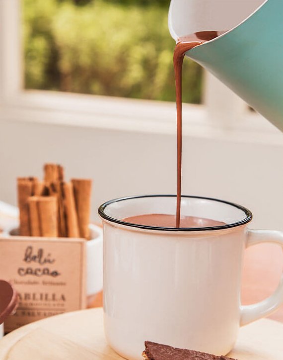 Chocolate is poured into a white mug with cinnamon sticks in the background.