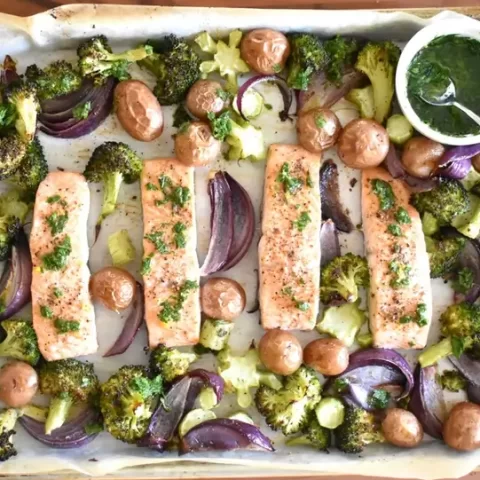 A baking sheet with salmon arranged over potatoes and drizzled with green sauce.