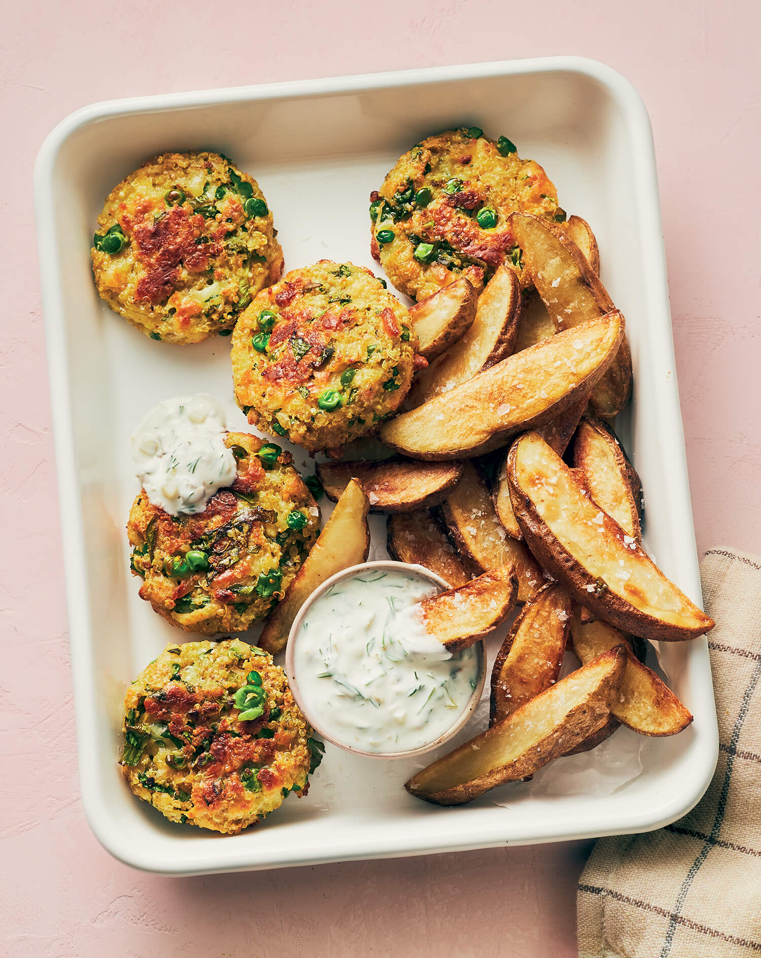 A tray with pea fritters, potato wedges and tartar sauce on a light pink background