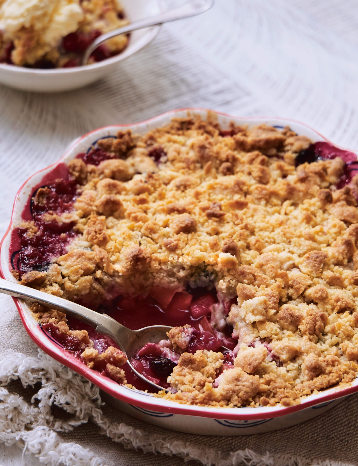 A rhubarb crumble with one serving in a white bowl on white linen