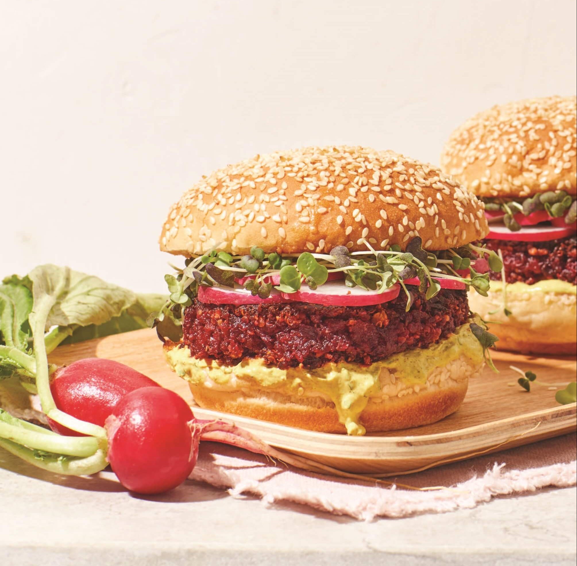Two veggies burgers with a bunch of beets next to them against a pale pink background
