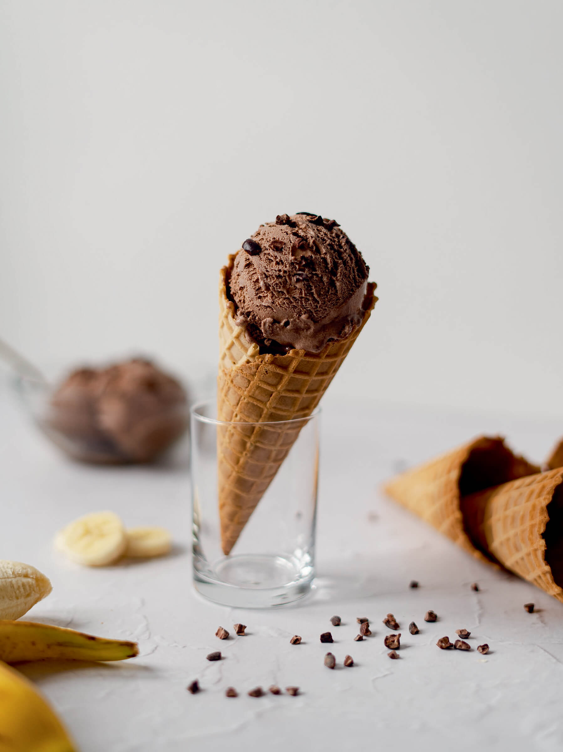 A chocolate ice cream cone resting in a glass with sliced bananas and other ingredients scattered nearby