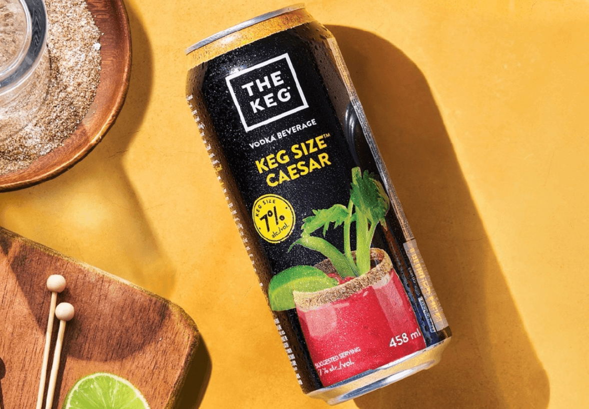 A canned Caesar cocktail on a yellow background with wooden boards with rim salt and lime nearby.