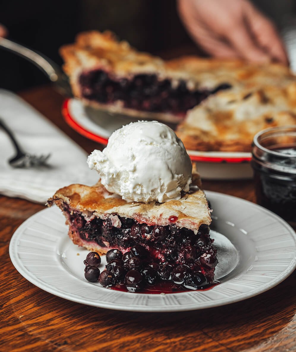 A slice of Saskatoon berry pie with a scoop of ice cream on top and a person serving more slices in the background