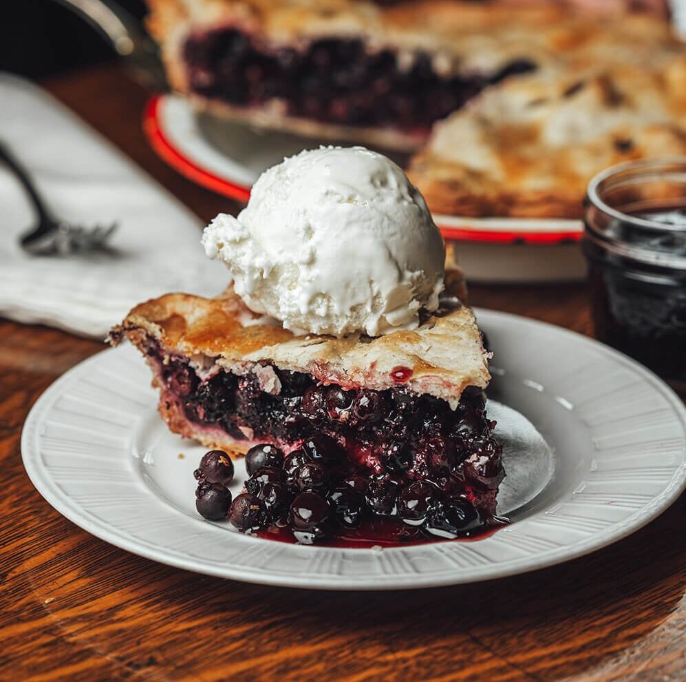 A slice of Saskatoon berry pie with a scoop of ice cream on top and a person serving more slices in the background