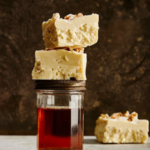 A jar of maple syrup with squares of white fudge stacked on top and one in the background.