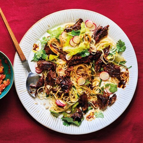 A white plate with rice noodle salad and short ribs on a bright red surface