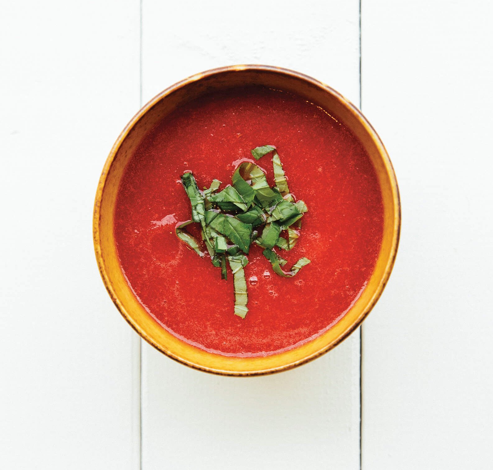 A small bowl of bright red soup with a lettuce garnish on bright white planks