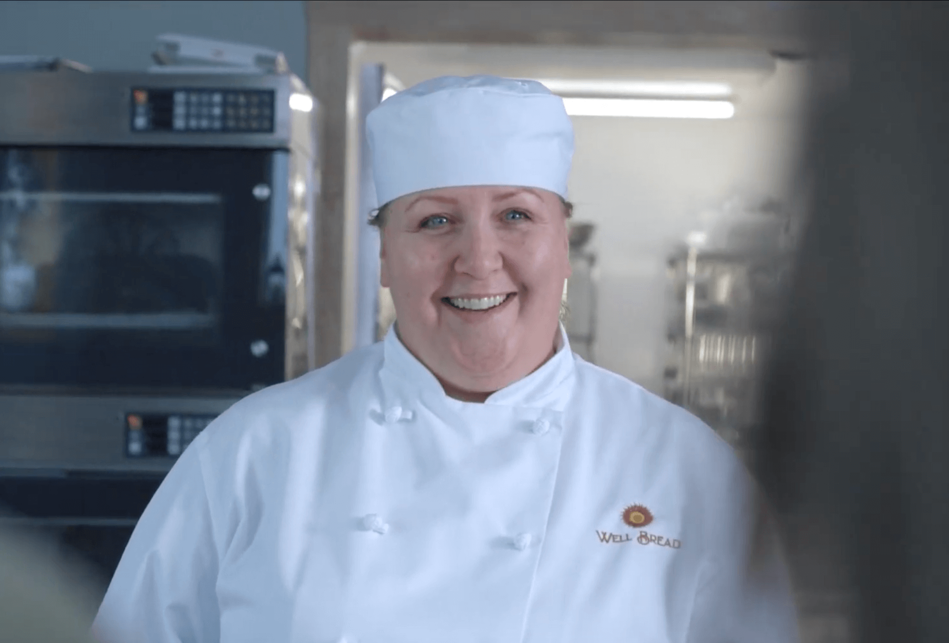 A woman in white chefs' attire smiling in an industrial kitchen