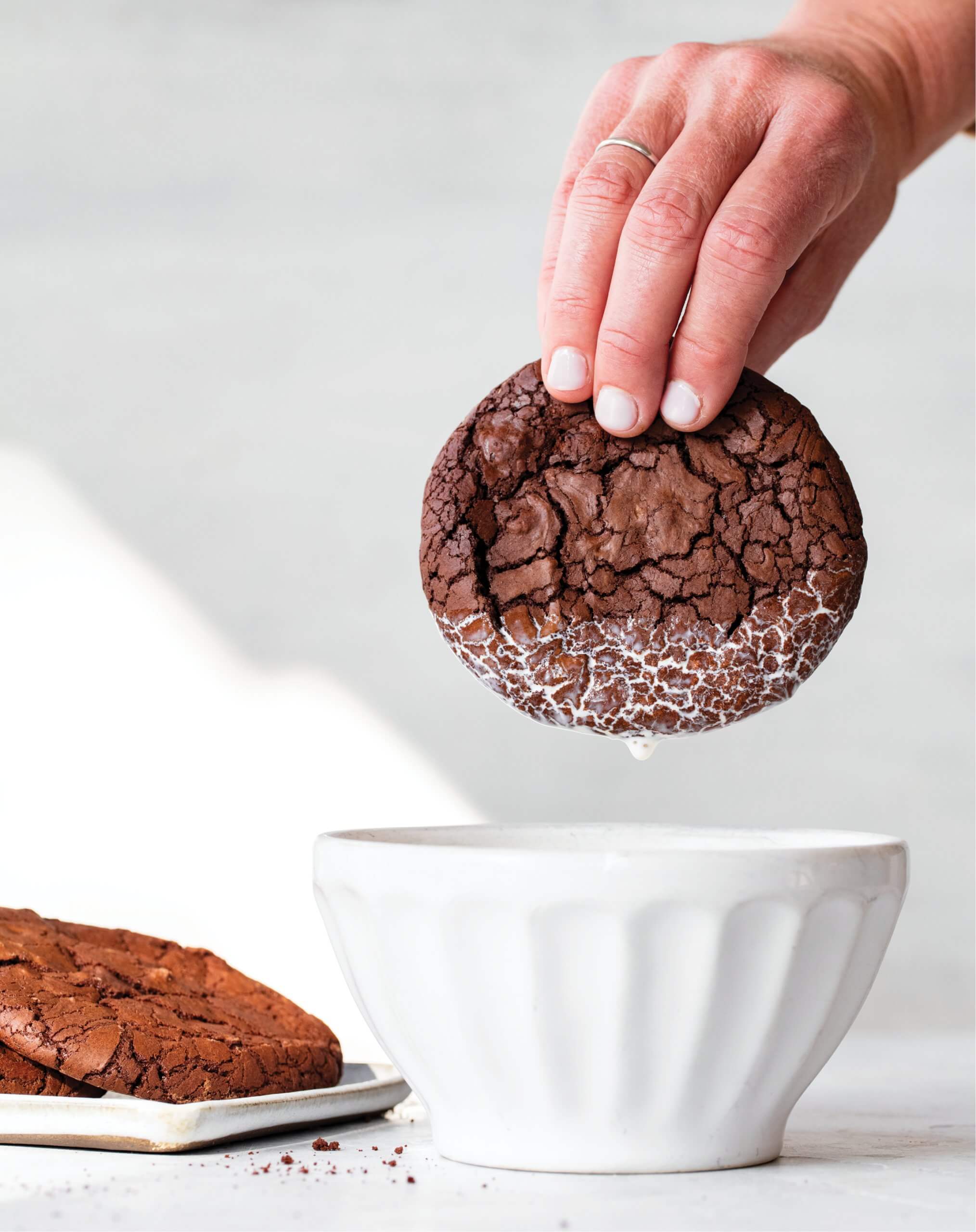 A person's hand dunking a chocolate cookie in a white bowl