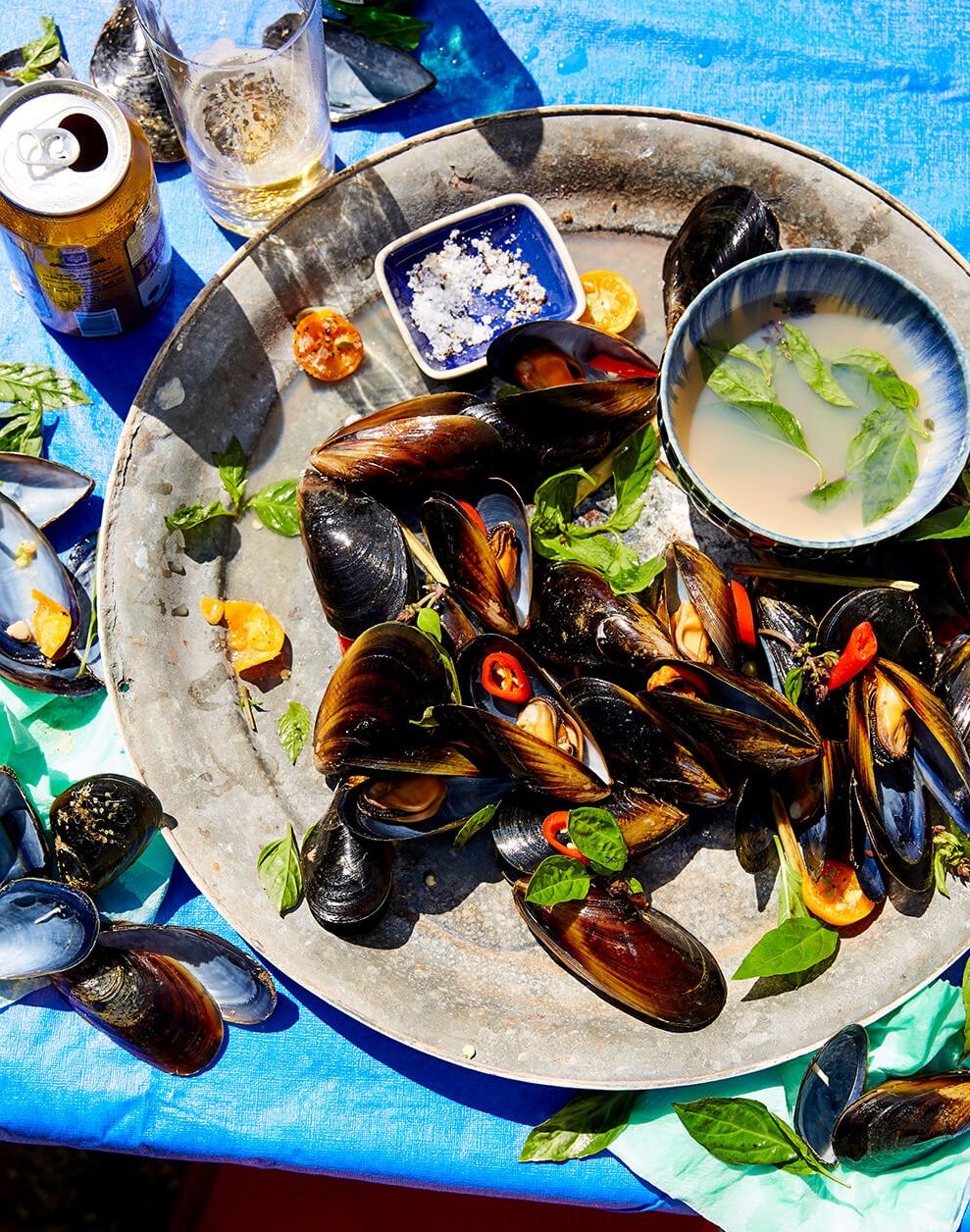 A plate of mussels on a bright blue tabletop, with salt and broth served on the side