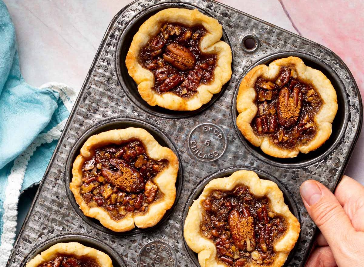 A person's hand holding tray of butter tarts over a blue and pink background