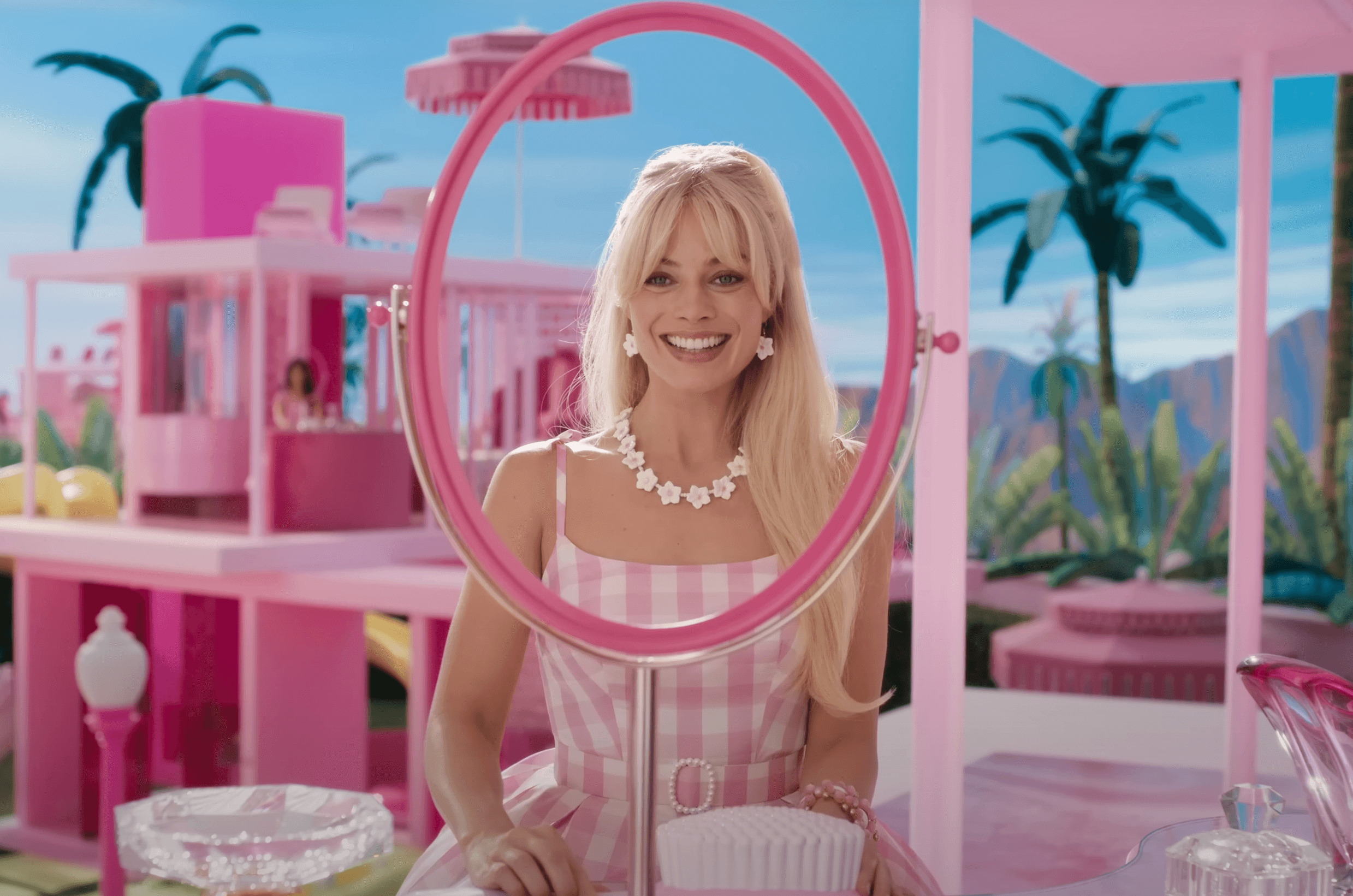 Margot Robbie as Barbie stands at a pink table with the outline of a mirror around her face