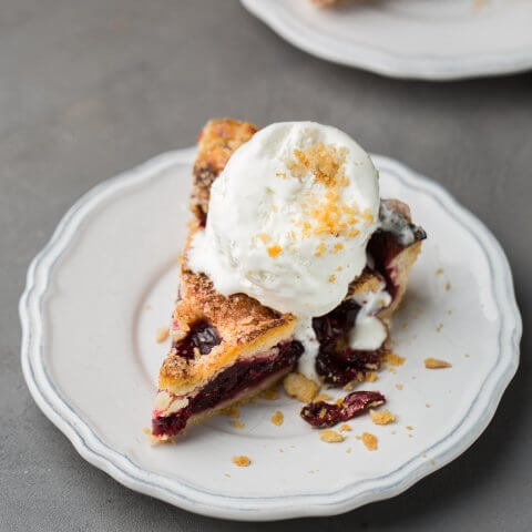 A slice of cherry pie on a white plate with a scoop of vanilla ice cream on top