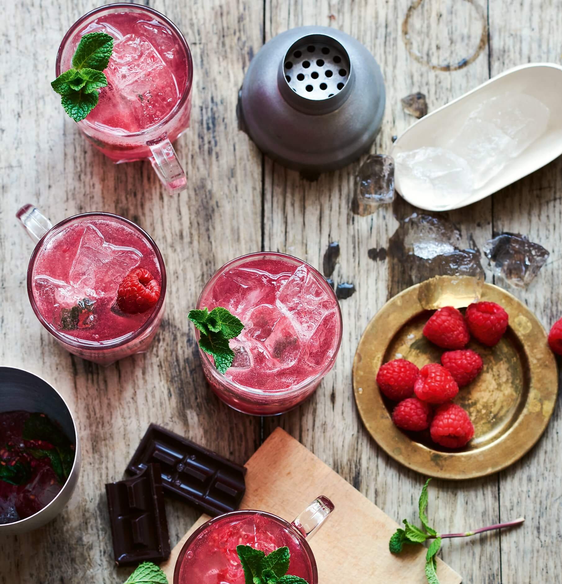 Glasses filled with a red cocktail and raspberries on a grey wooden surface