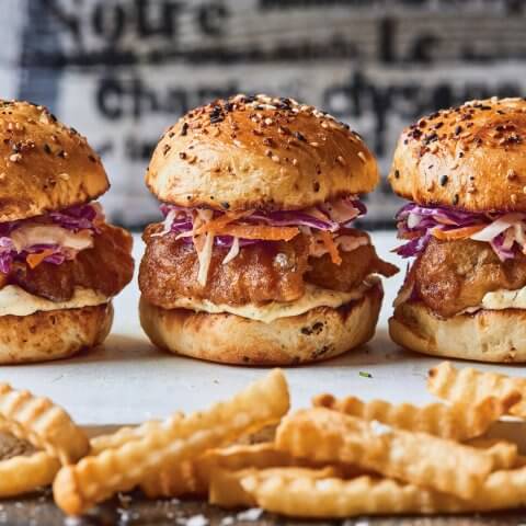 Three fried fish sandwiches and crinkle-cut fries on a white surface with black and white type visible on the wall behind it