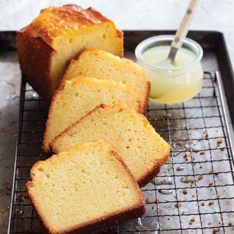 A sliced yellow lemon loaf cake on a drying rack, with a container of lemon drizzle next to it