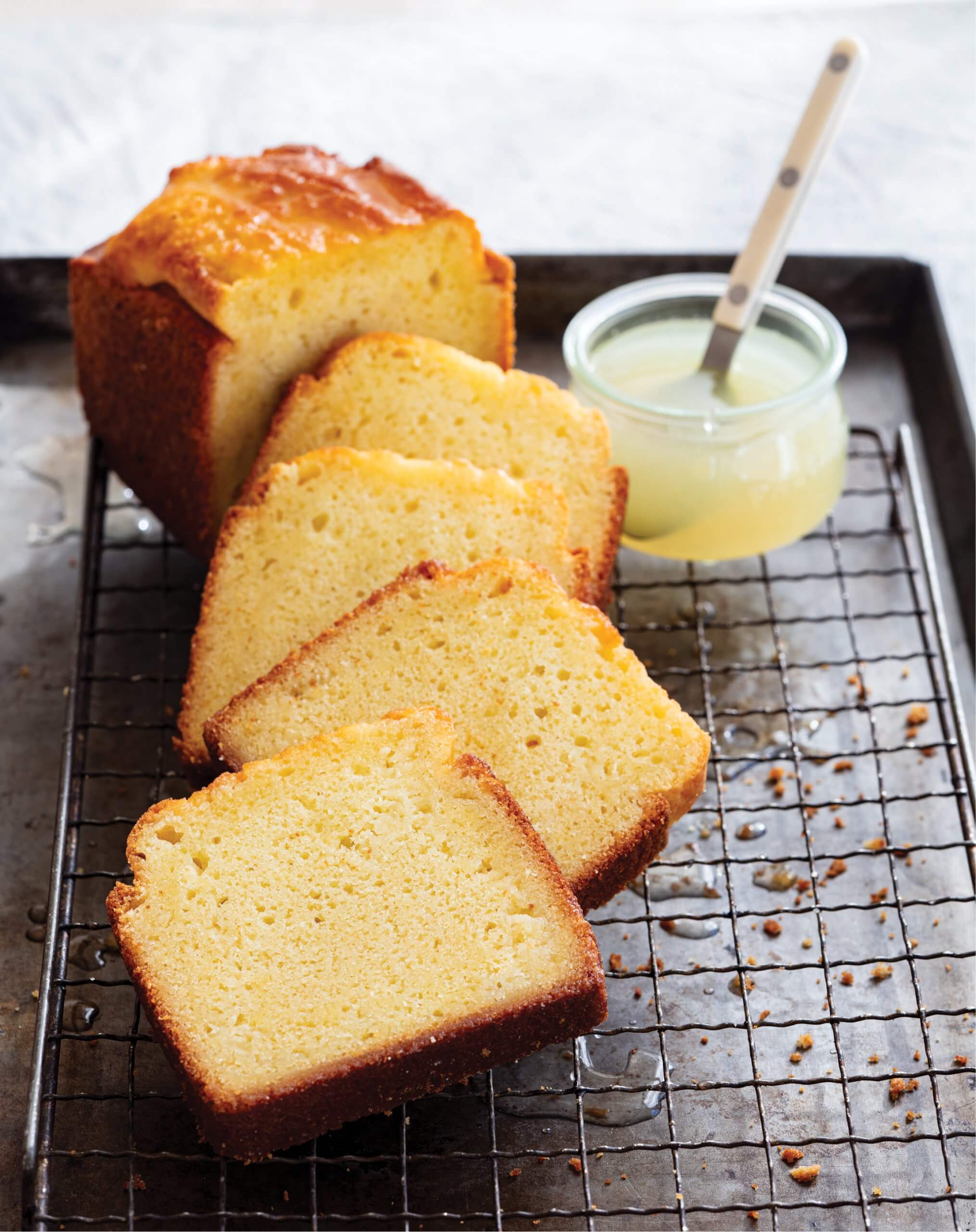A sliced yellow lemon loaf cake on a drying rack, with a container of lemon drizzle next to it