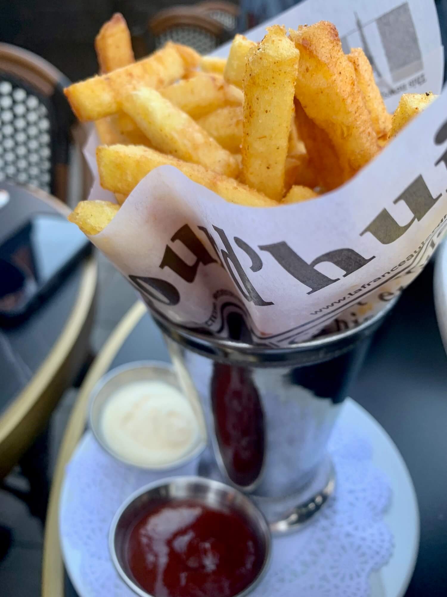 A metal cone with french fries wrapped in paper and ketchup and mayonnaise on the side