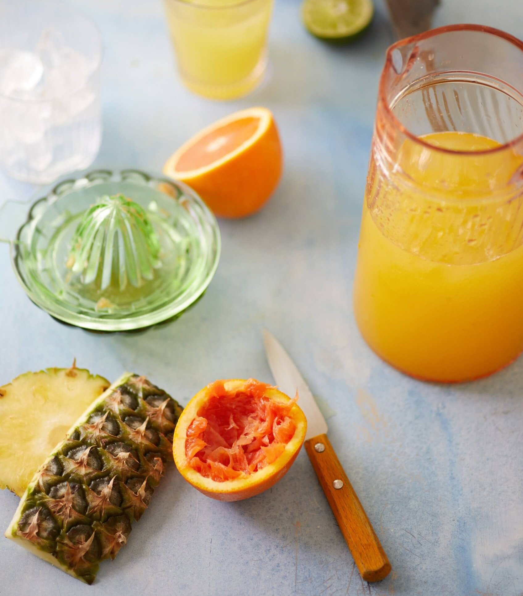 Sliced oranges, pineapples and limes on a white surface with a green glass juicer and pitcher of juice nearby