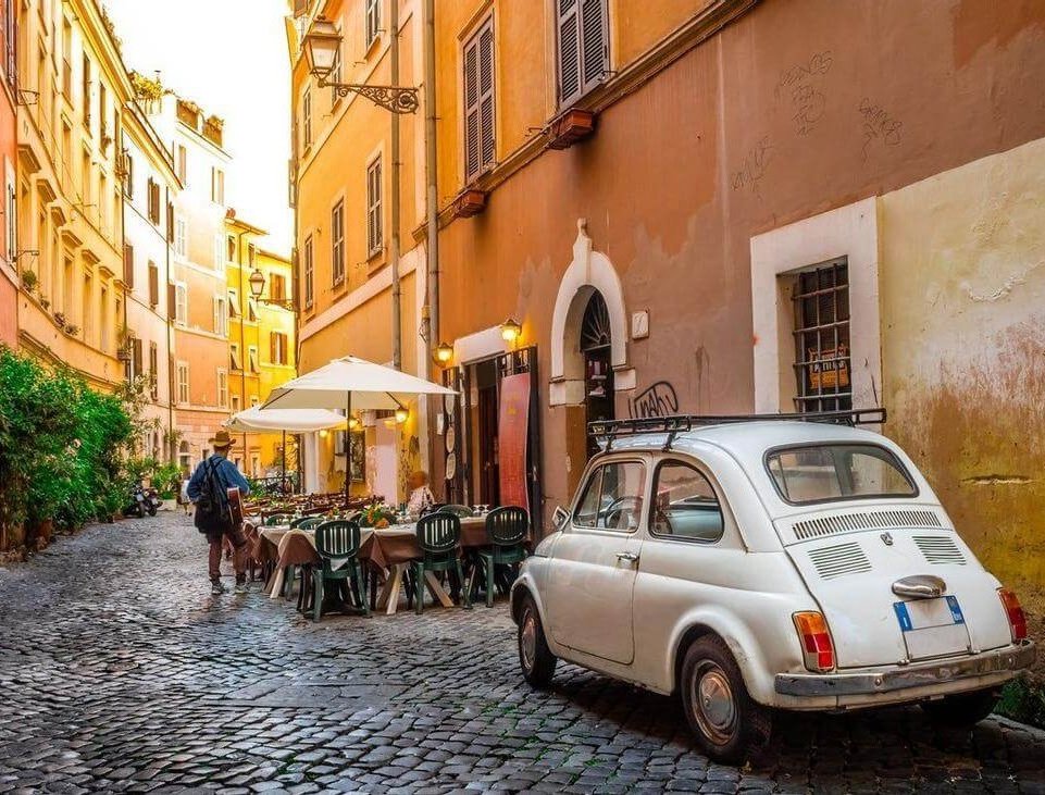 A view of a cobbled street in Rome, with an outdoor cafe and a white Fiat.