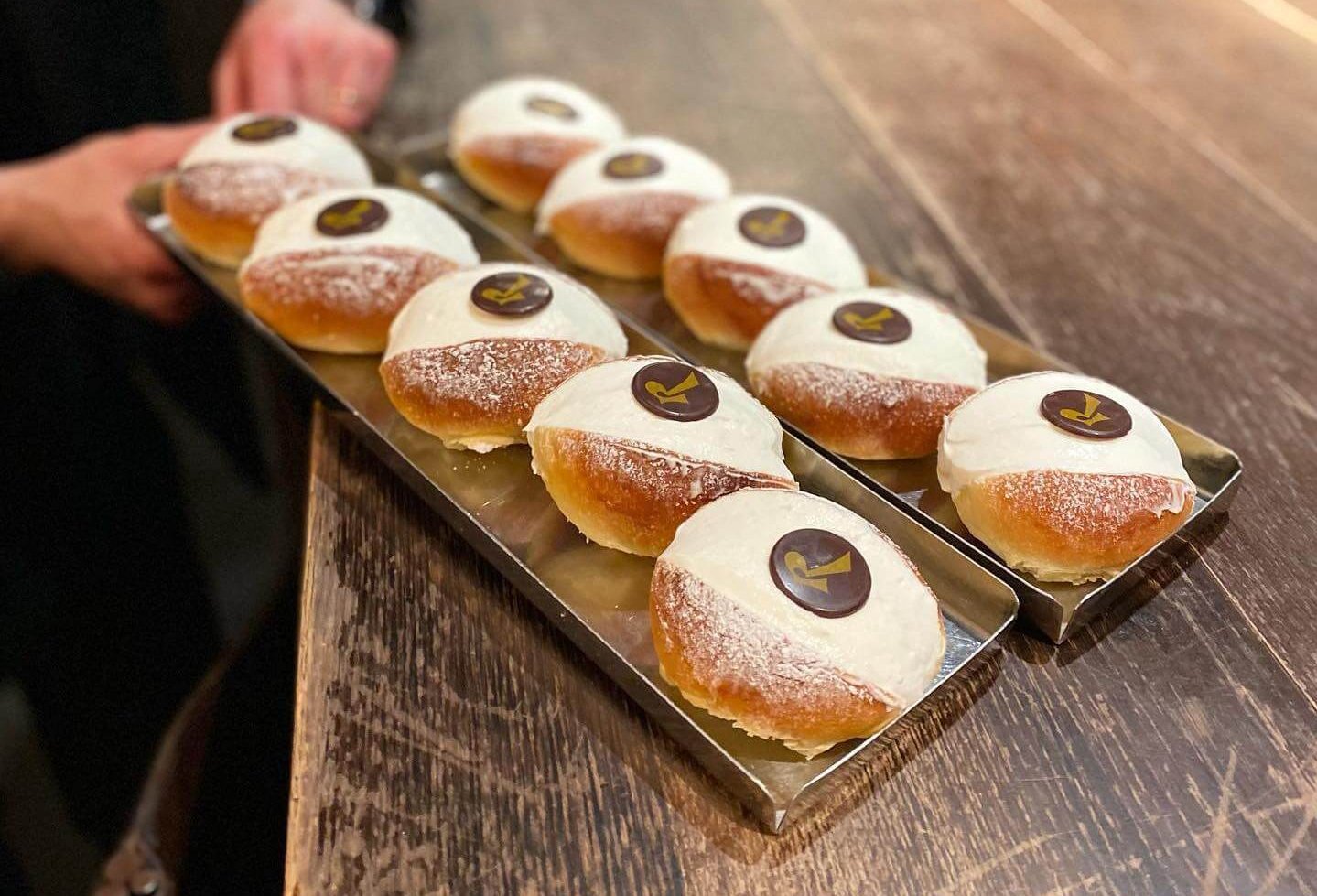A person holding silver trays of cream buns topped with a chocolate circle bearing a gold 'R' over a wooden tabletop