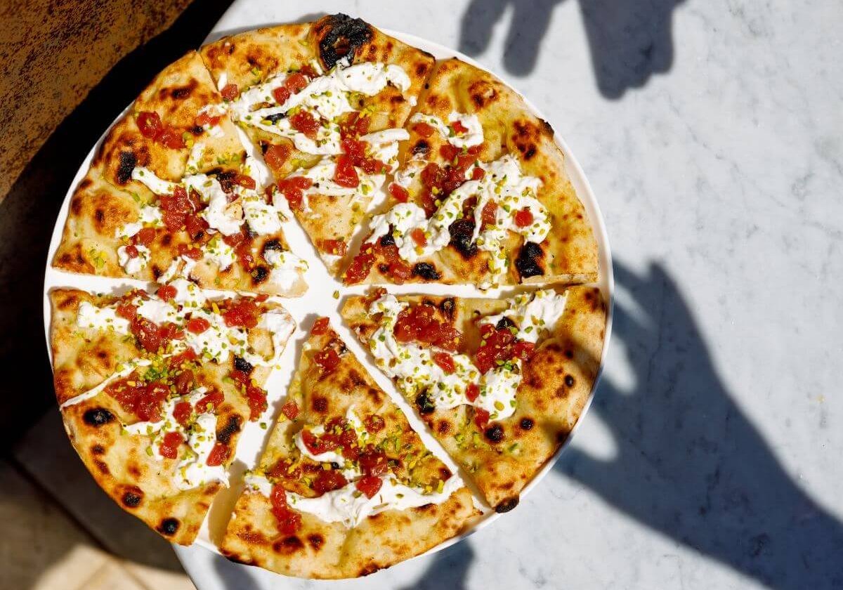 A pizza on a marble tabletop with the shadows of peoples' hands reaching for it