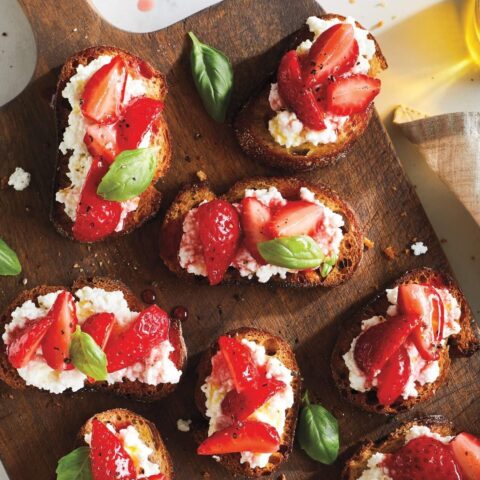 A dark wood serving board laid crostini, goat cheese and strawberry appetizers and a sprinkling of fresh basil leaves