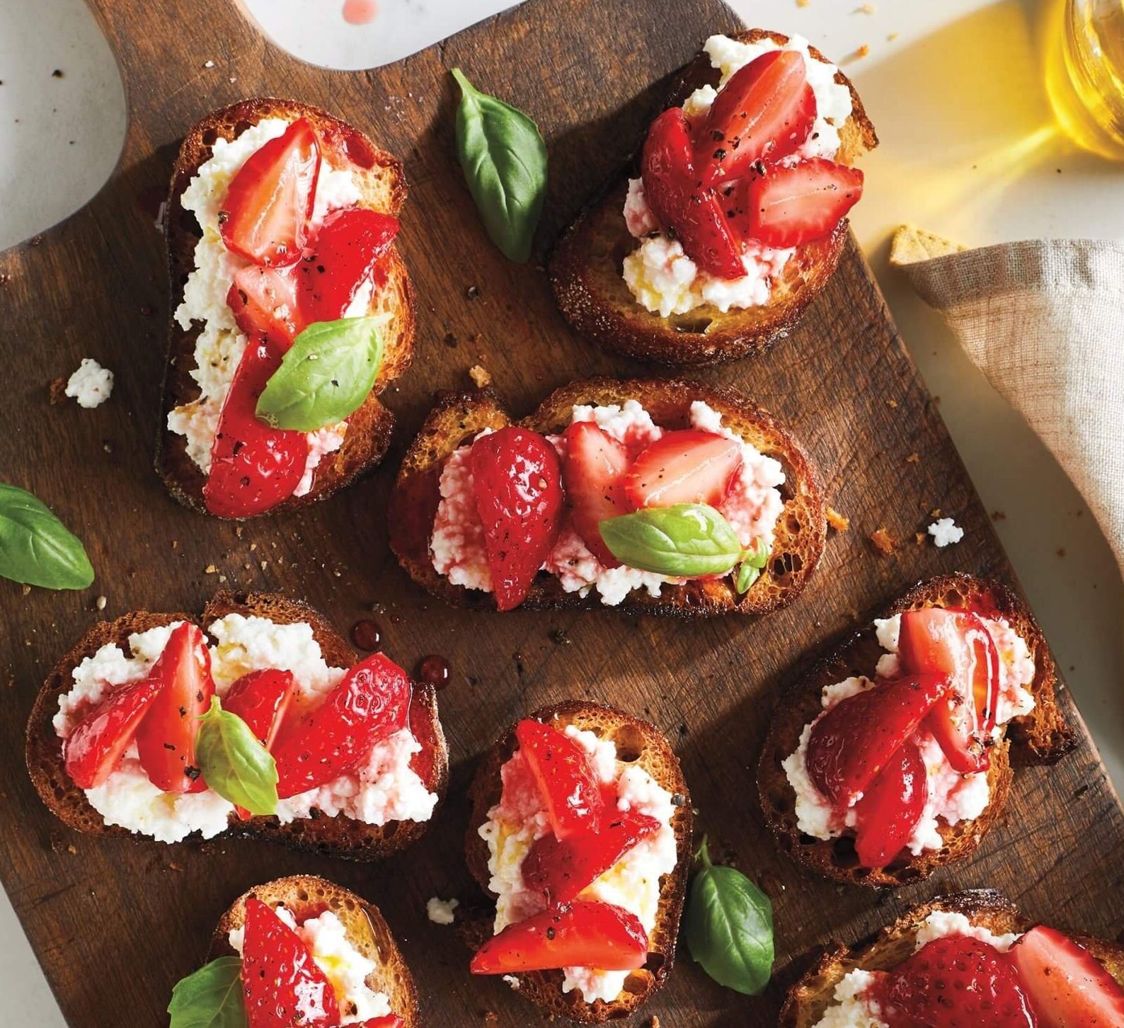 A dark wood serving board laid crostini, goat cheese and strawberry appetizers and a sprinkling of fresh basil leaves